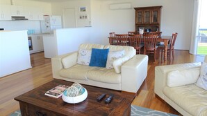 A modern living room with a beige sofa, a coffee table with magazines and a decorative item, a dining area with wooden chairs and a table, and a kitchen with white cabinets and appliances. - Shell Seeker, Moonta Bay (Moonta Bay)