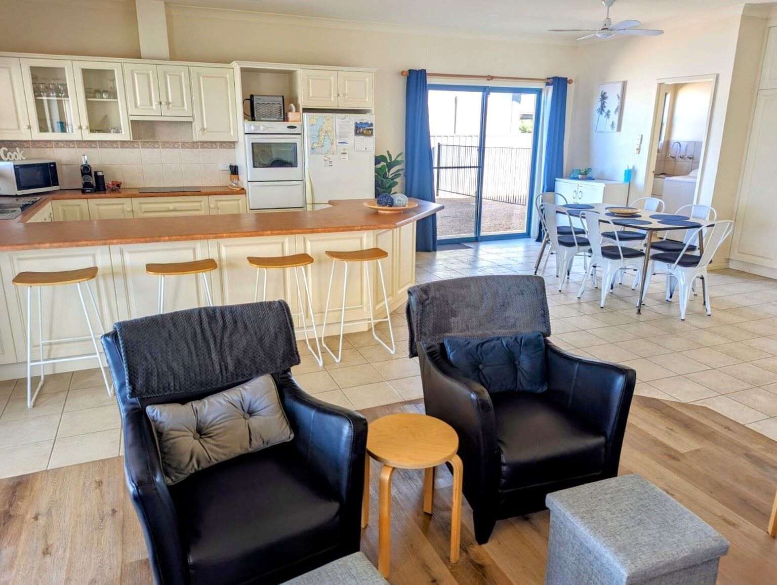A modern kitchen with white cabinets and a central island, a dining area with white chairs, and two black leather sofas with cushions.
