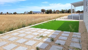 A paved patio with a green lawn area, a white building with a covered porch, and a field in the background.