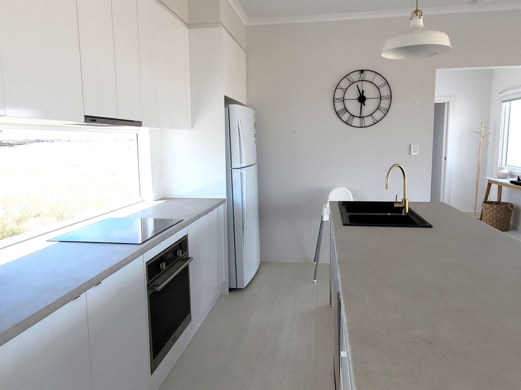 A modern kitchen with white cabinetry, a stainless steel refrigerator, a built-in oven, a black countertop, a white sink with a gold faucet, and a wall-mounted clock.