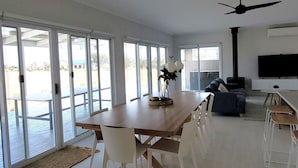 A spacious living area with a large wooden dining table, white chairs, a sofa, and a television.