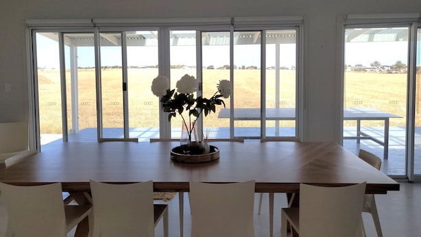 A dining area with a large table and white chairs, large windows with a view of a field, and a vase with flowers on the table.