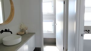 A bathroom with a large round mirror, a white sink, and a window with blinds.