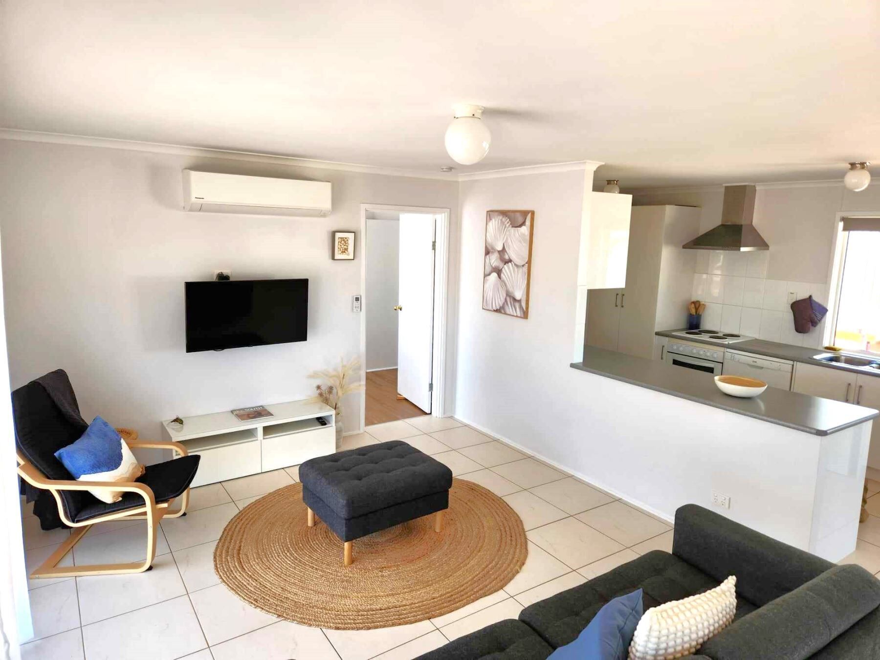 Ein modernes Wohnzimmer mit Fernseher, Sofa und Hocker, eine Küche mit Herd und Schränken sowie ein Essbereich mit Tisch und Stühlen.