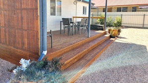 Een overdekt terras buiten met houten vloer, een tafel, stoelen en potplanten.