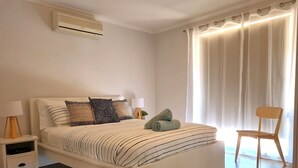 Een netjes ingerichte slaapkamer met een bed, nachtkastjes, een lamp, een stoel en een raam met gordijnen.