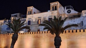 Un bâtiment historique doté d’arcs en plein cintre et d’un mur fortifié, avec des palmiers en premier plan.