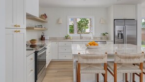 Una cocina moderna con armarios blancos, encimera de mármol y refrigerador de acero inoxidable. Se ve vegetación por la ventana.