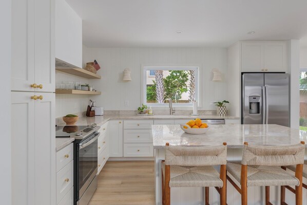 A modern kitchen with white cabinetry, a marble countertop, and a stainless steel refrigerator. There is a window with a view of greenery outside.
