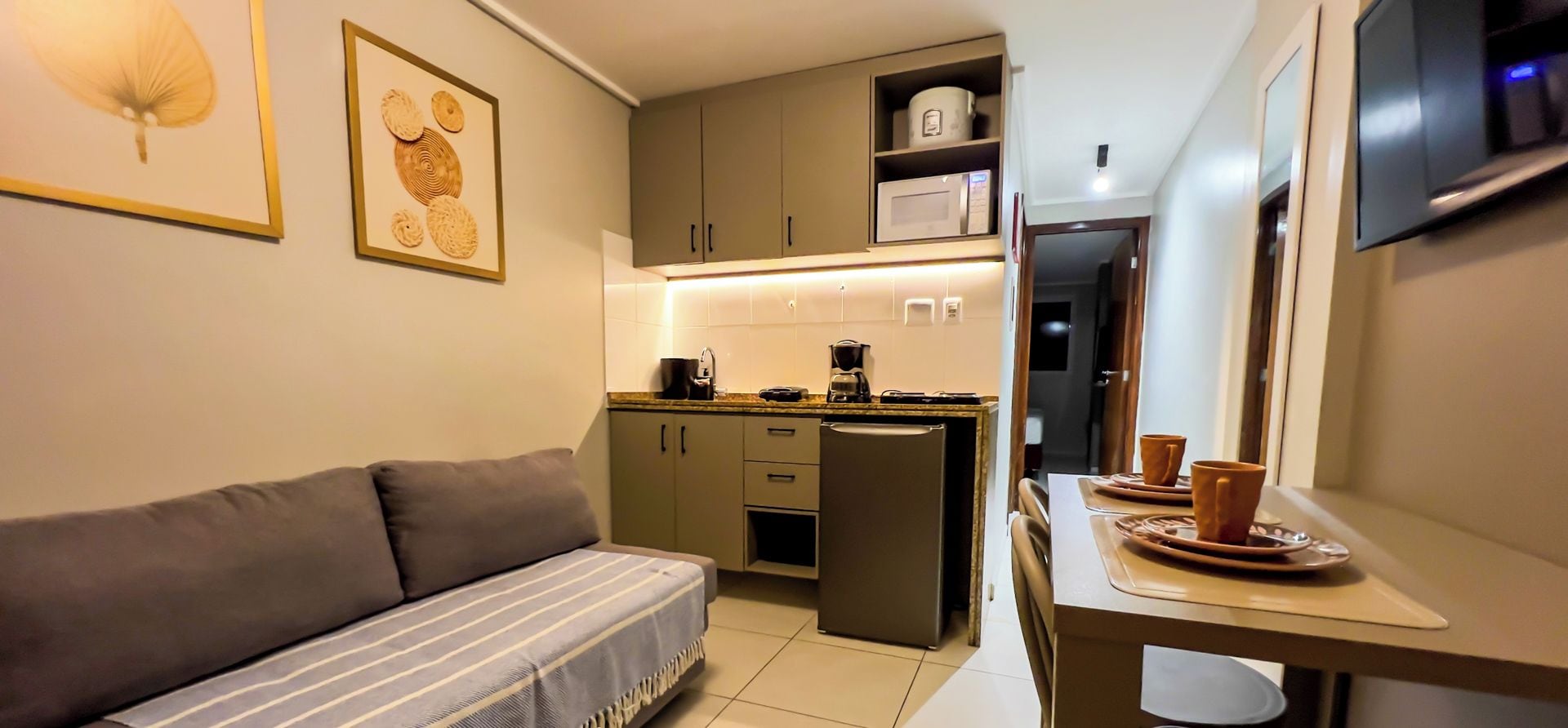 Espaço compacto com sofá, mesa de jantar com cadeiras, cozinha com armários e eletrodomésticos, e televisão fixada na parede.