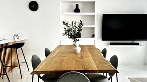 A modern dining area with a wooden table, black chairs, a white vase with plants, a black clock, and a TV on a white shelf.