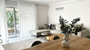 A modern living room with a sofa, a coffee table, and a dining table. There is a sliding glass door leading to a balcony with plants.