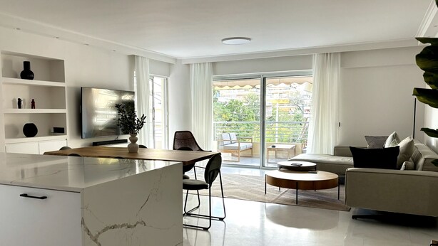 A modern living room with a dining area, a sofa, a TV, and a balcony with a view of greenery.