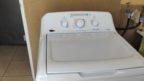 A front-loading washing machine with control knobs and a digital display, located in a room with tiled flooring and a visible water pipe. - Hidden Gem, Cozy 2-bedroom house with WiFi, AC, Private Pool In Portmore (Portmore)