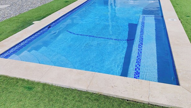 A rectangular swimming pool with a tiled edge, surrounded by a grassy area and a white wall. - Hidden Gem, Cozy 2-bedroom house with WiFi, AC, Private Pool In Portmore (Portmore)