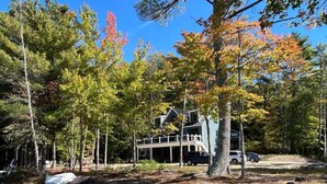 A two-story house with a deck, surrounded by trees and a dirt path. - The Beach House on Shaws Pond - gorgeous new home directly on the water (New Durham)