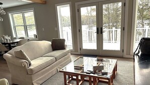 A living room with a beige sofa, a glass coffee table, and a view through large glass doors to an outdoor deck. - The Beach House on Shaws Pond - gorgeous new home directly on the water (New Durham)