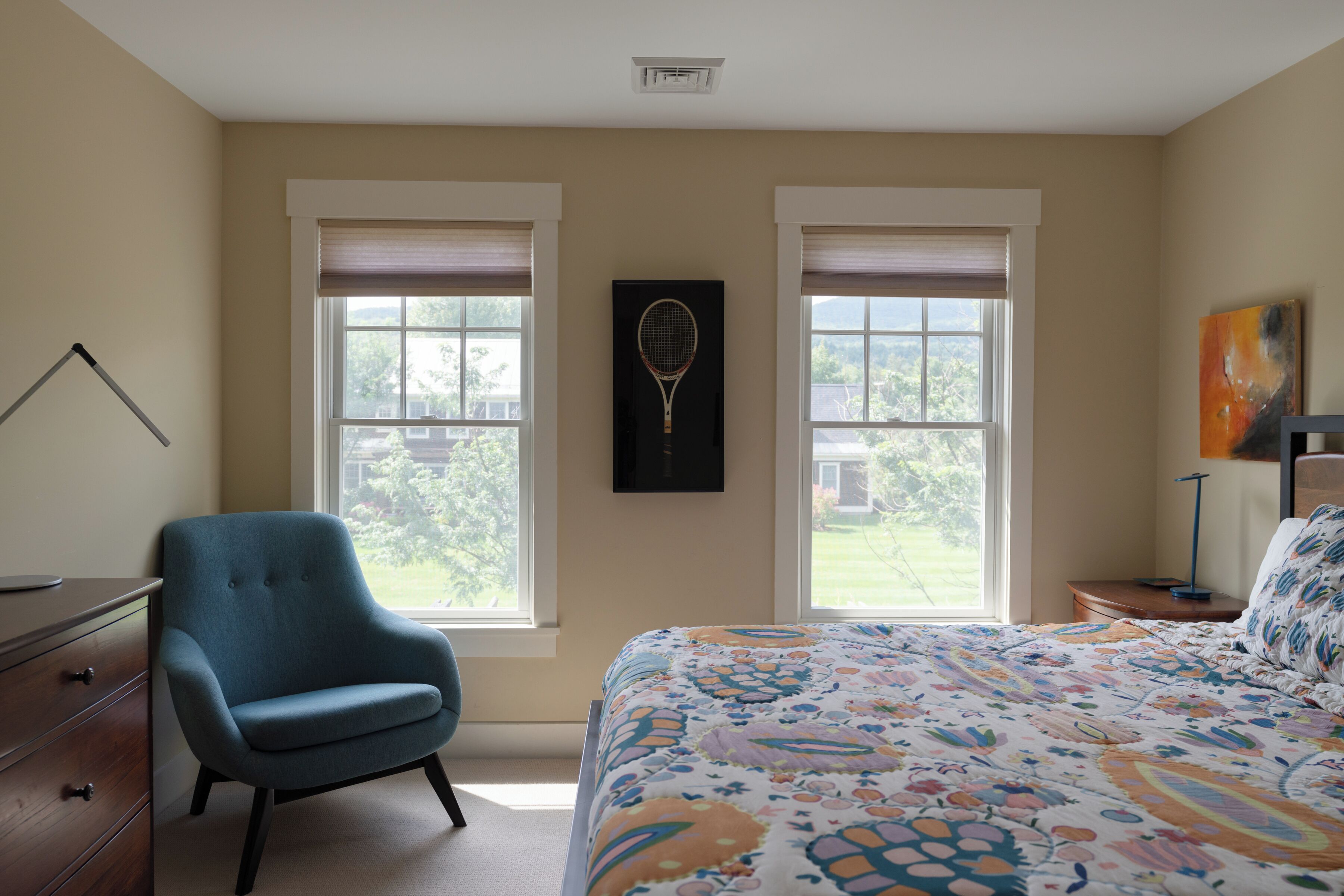 A bedroom with a bed, a chair, a desk, and two windows with blinds.