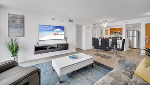 A modern living room with a flat-screen TV, a white coffee table, and a grey sofa. There is a dining area with a table and chairs, and a kitchen with a refrigerator and microwave. - Amazing spacious unit (Miami)