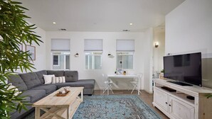 Un salon moderne avec un canapé gris, une table basse en bois, un téléviseur posé sur un meuble blanc et un coin repas avec une table et des chaises blanches.