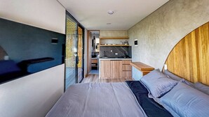 Ein modernes, kompaktes Schlafzimmer mit einem großen Bett, einer kleinen Küchenzeile und einer Glastür, die auf einen Balkon führt.