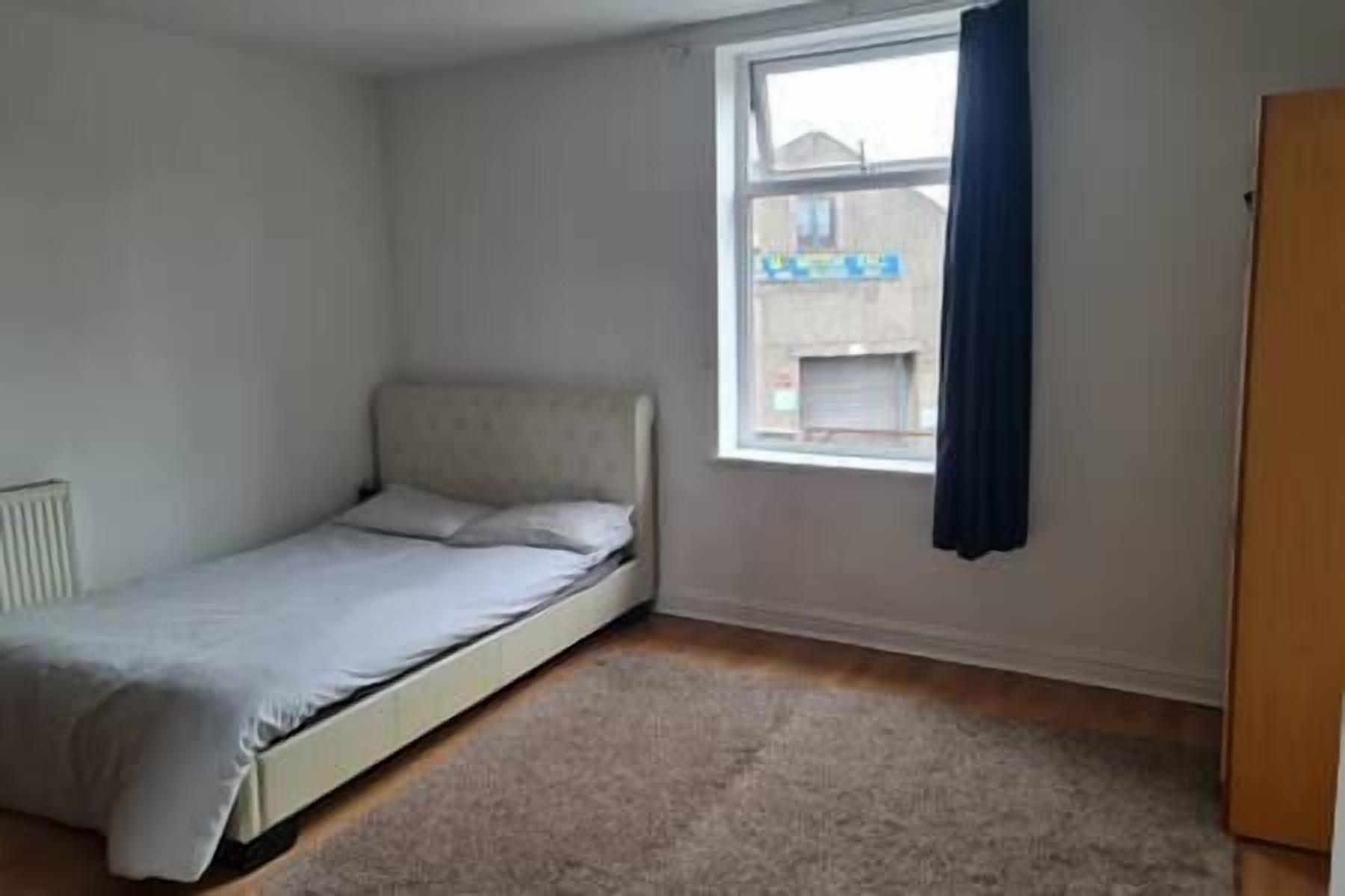 Een kleine, eenvoudig ingerichte kamer met een enkel bed, een radiator, een raam met gordijnen en een deur.