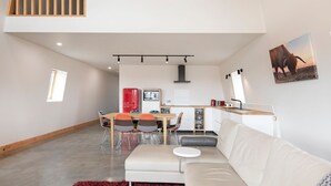 A modern living room with a white sofa, a dining table with chairs, a kitchen area with a red refrigerator, and a large window.