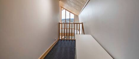 A narrow stairway with a wooden railing, carpeted steps, and a view of a landscape through a large window at the top.