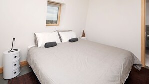 A neatly made bed with a grey bedspread and two pillows, a small bedside table with a statue, a window with a view of trees, and a white storage unit with drawers.