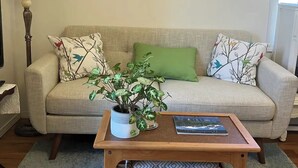 En stue med sofa, to puder med mønster, en grøn pude, et træ sofabord med en plante og en bog, en gulvlampe og en dekorativ vægdekoration.