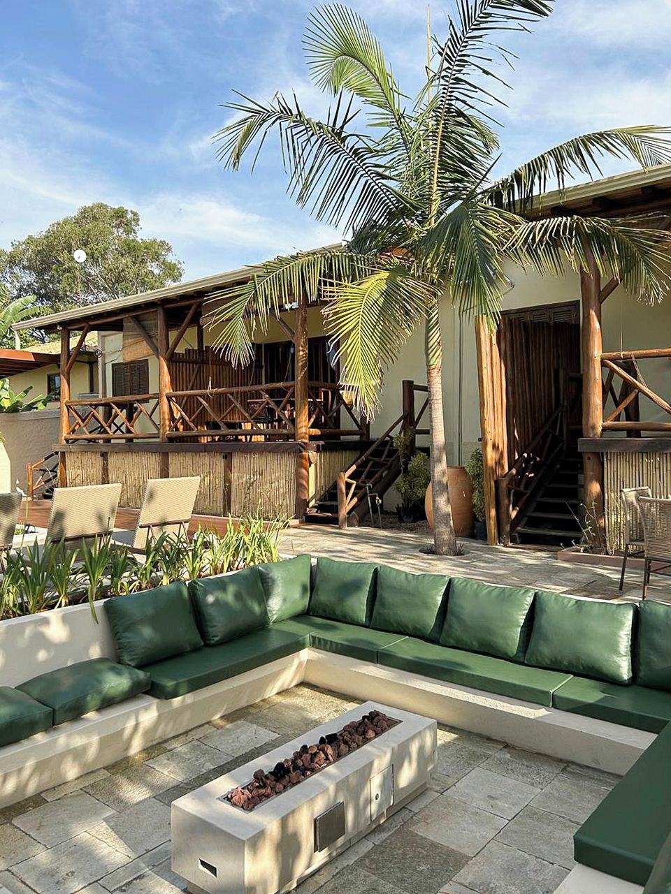 Un espace patio aménagé avec un canapé vert, un foyer extérieur et une terrasse en bois dotée d’un escalier menant à une zone abritée.