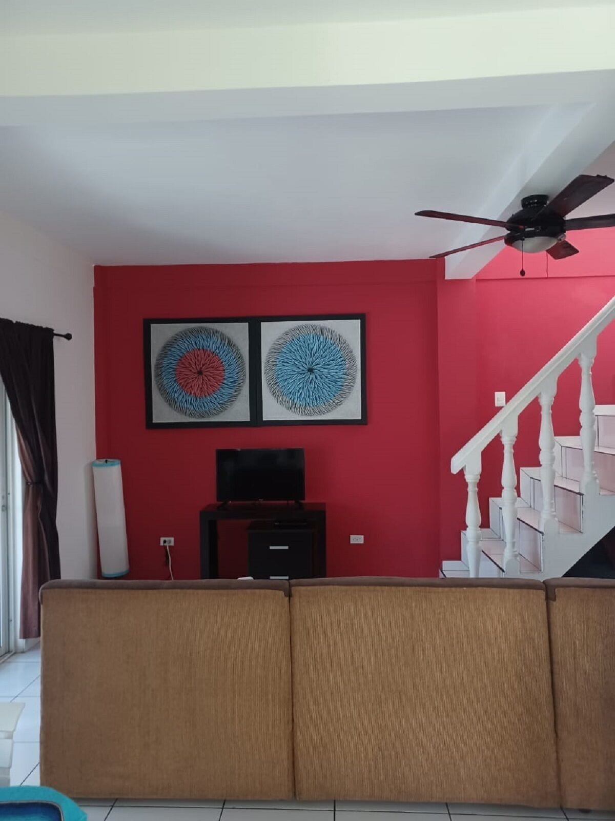 Una sala de estar con una pared roja, dos piezas de arte circular en la pared, un ventilador de techo, una escalera y un sofá marrón.