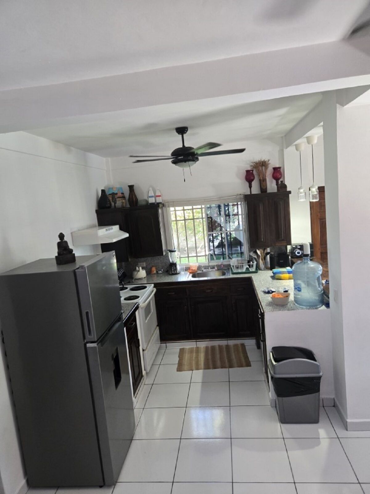 A kitchen with a refrigerator, stove, and sink. There is a ceiling fan, a window with a view, and a water dispenser.