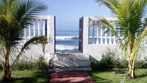 Un sendero que lleva a una playa con vista al océano, bordeado por palmeras y un edificio blanco con balcón.