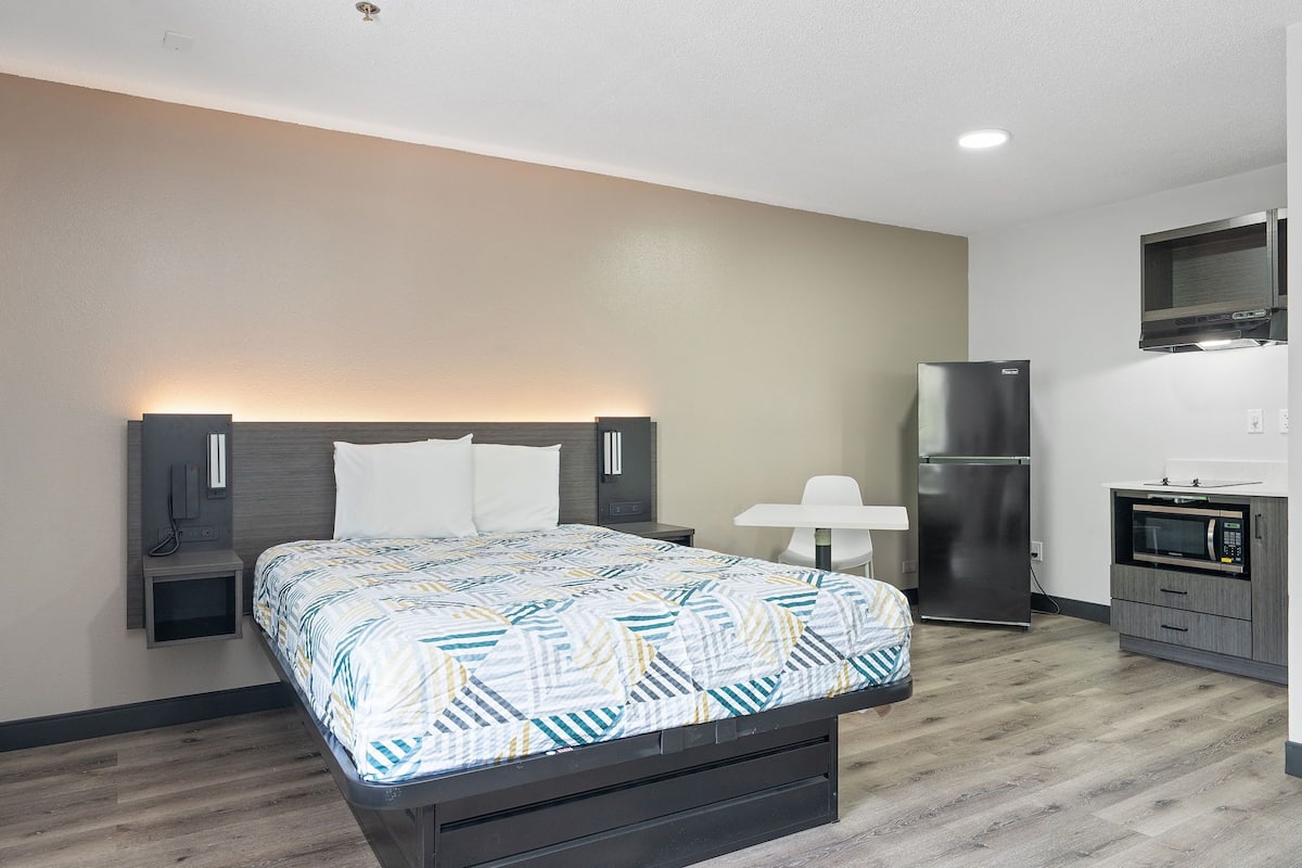 A modern hotel room with a bed, bedside tables, a small desk, a refrigerator, and a microwave.