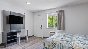 Ett hotellrum med en säng, ett skrivbord med en stol, en platt-TV, ett fönster med gardiner och en dörr.