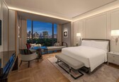 A modern hotel room with a large bed, a desk, and a view of a cityscape.