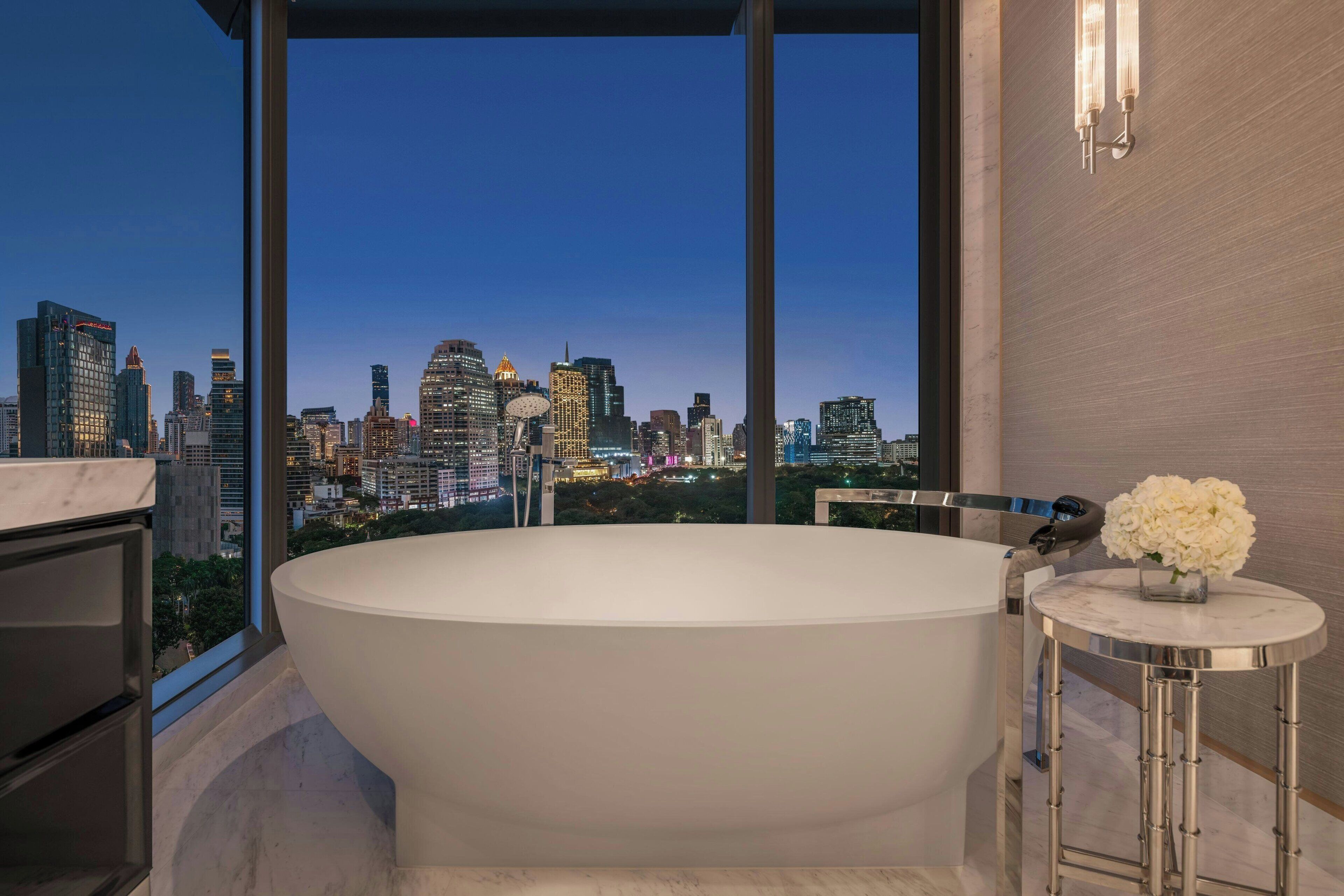A modern bathroom with a freestanding bathtub, a marble vanity, and a view of a city skyline at dusk.