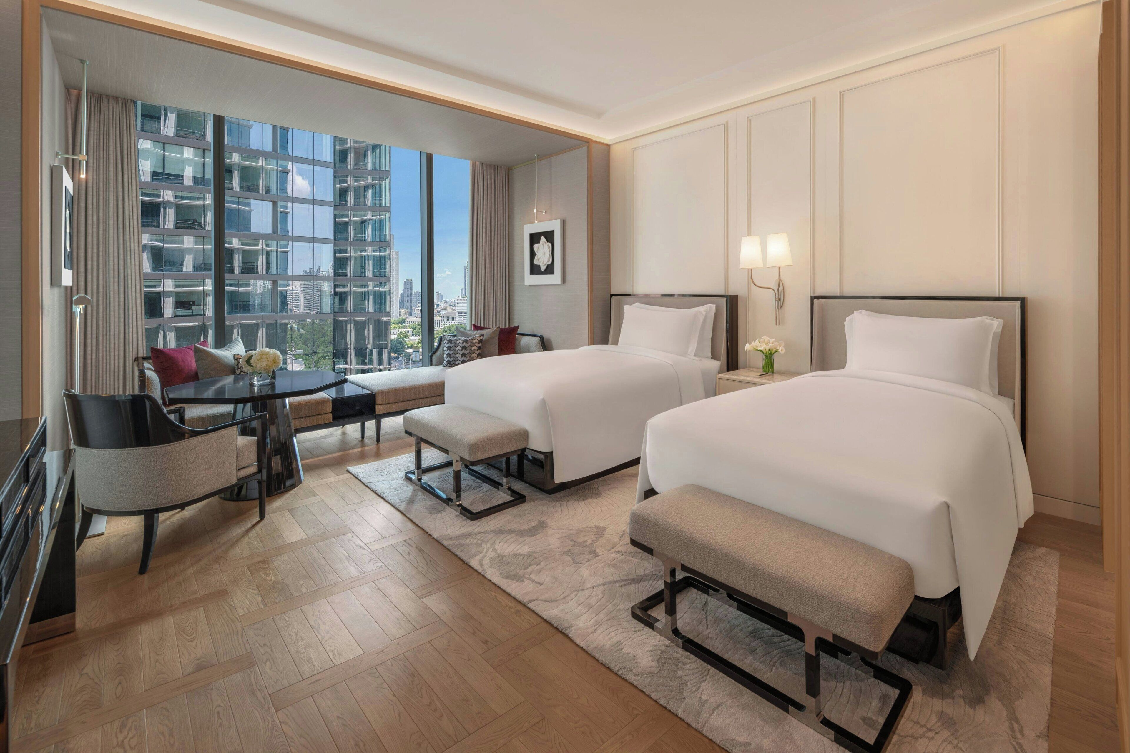 Habitación de hotel con dos camas, un escritorio, una silla y vistas a la ciudad a través de amplias ventanas.