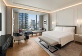 A modern hotel room with a large bed, bedside tables, a desk with a chair, a sofa, and a view of the cityscape.