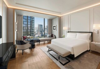 A modern hotel room with a large bed, bedside tables, a desk with a chair, a sofa, and a view of the cityscape.