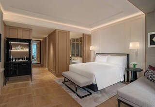 A hotel room with a large bed, a bedside table with a lamp, a bench, a wardrobe, and a mirror.