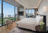 A modern hotel room with a large bed, a sitting area with a chair and table, a view of the cityscape, and a vase with flowers on a side table.