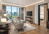 A modern hotel room with a large window offering a city view, a flat-screen TV, a seating area with a glass table, and a separate bedroom with a bed and a nightstand.