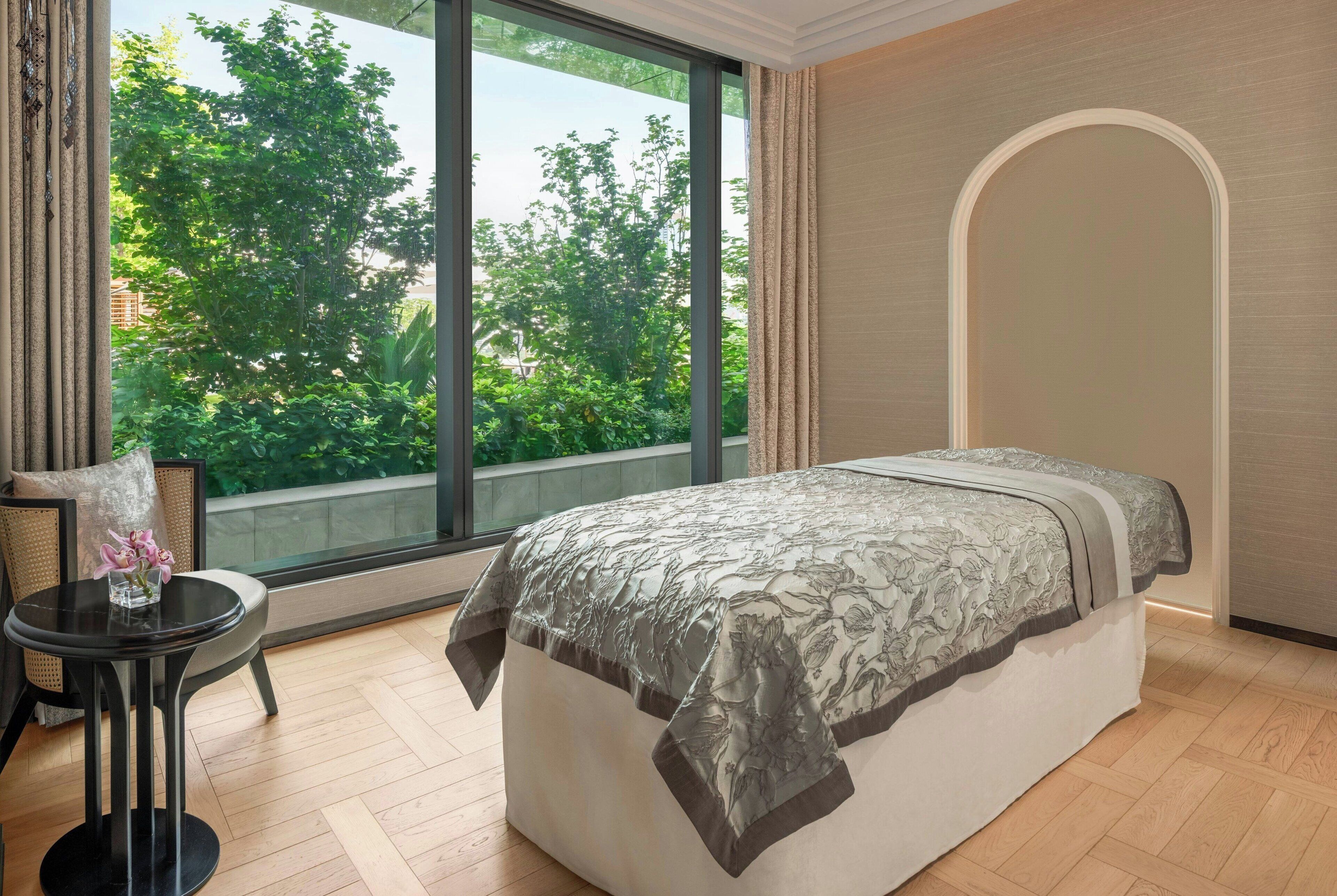 A bedroom with a large window, a bed with a floral bedspread, a small round table with a vase, and a chair.