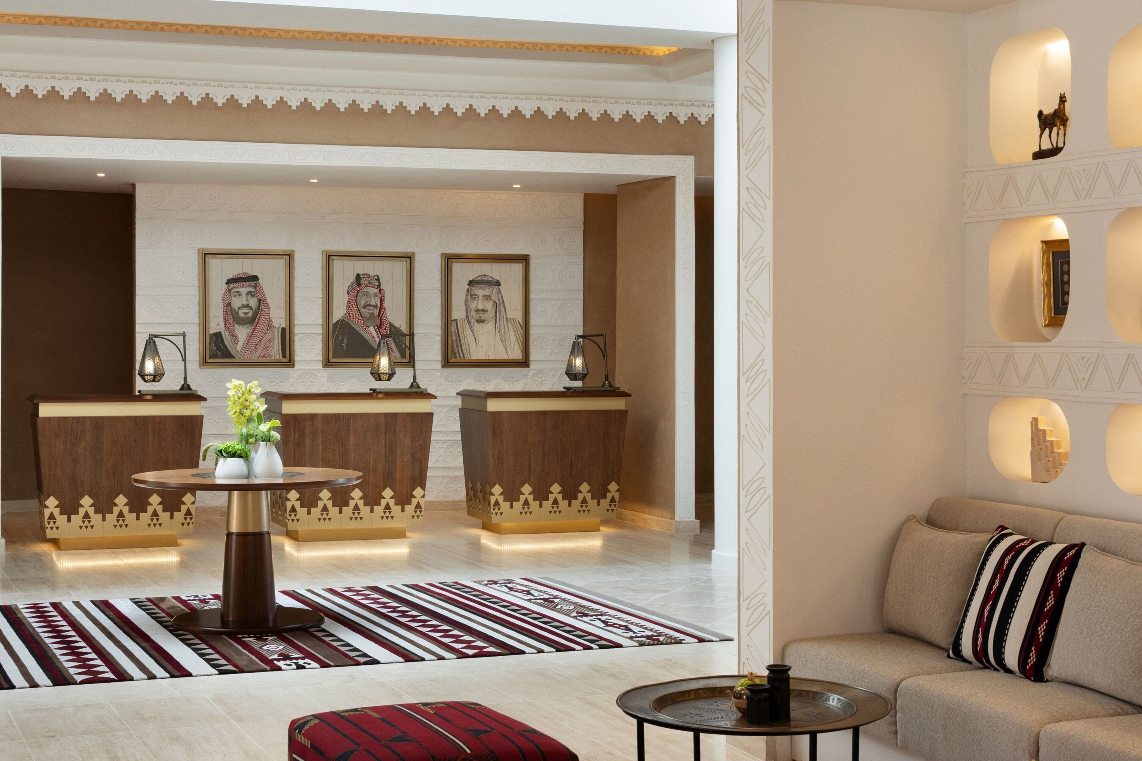 Un hall con mostrador de recepción, dos retratos enmarcados, una mesa redonda con un jarrón, un tapete estampado, un sofá con un cojín a rayas y una pared con iluminación decorativa.