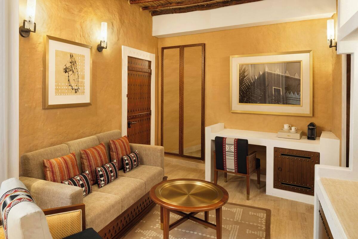 A cozy living area with a sofa, a coffee table, and a desk with a chair. There are framed pictures on the walls and a wooden cabinet.