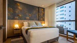 A modern hotel room with a large bed, two bedside tables with lamps, a view of a building through a large window, and a wooden floor.