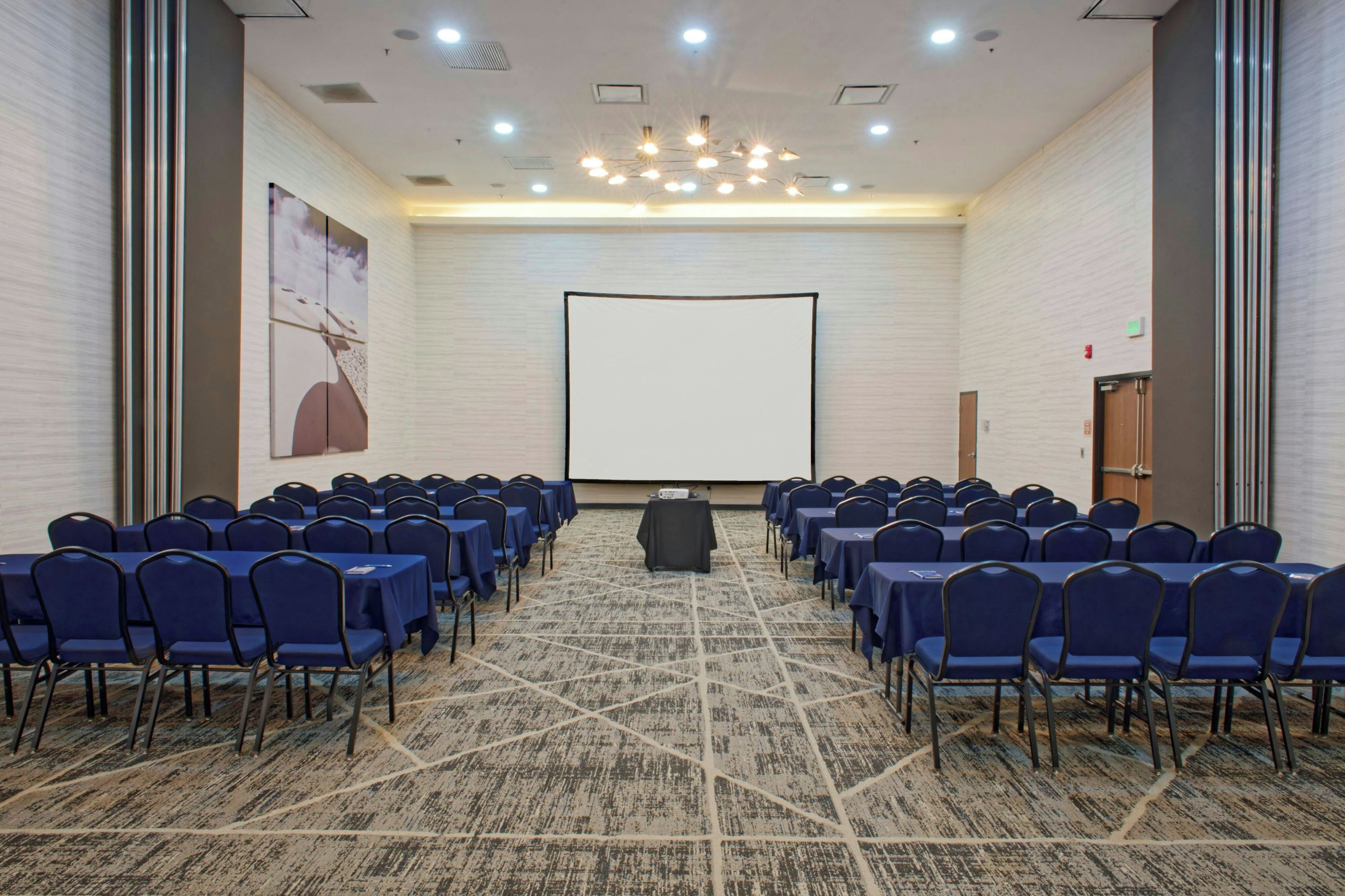 Ein Konferenzraum mit Reihen von Stühlen, die auf eine leere Projektionsleinwand zeigen, mit Teppichboden und einer großen Wand, auf der ein gerahmtes Bild hängt.