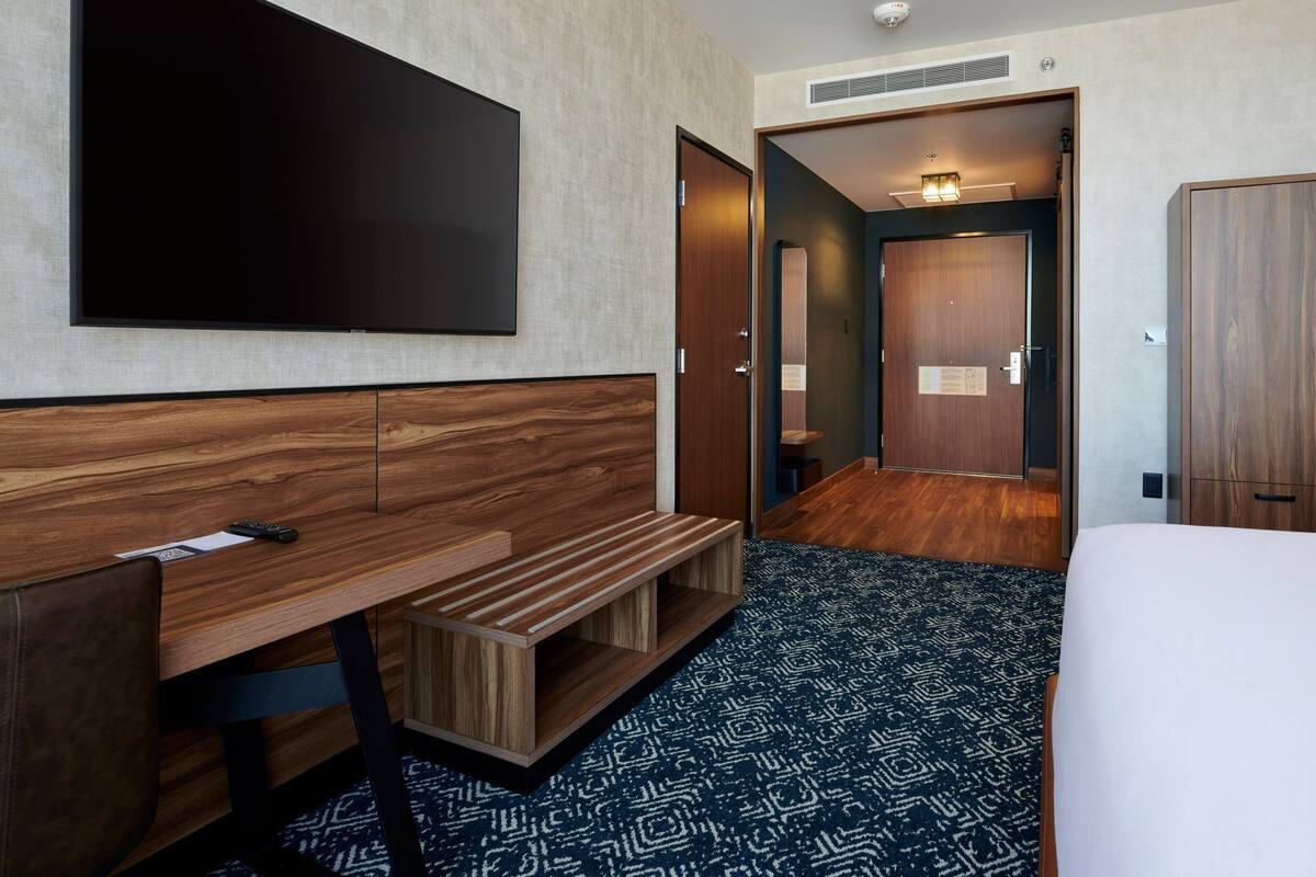 A hotel room with a large flat-screen TV mounted on the wall, a wooden desk with a bench, a bed with a patterned carpet, and a wardrobe.
